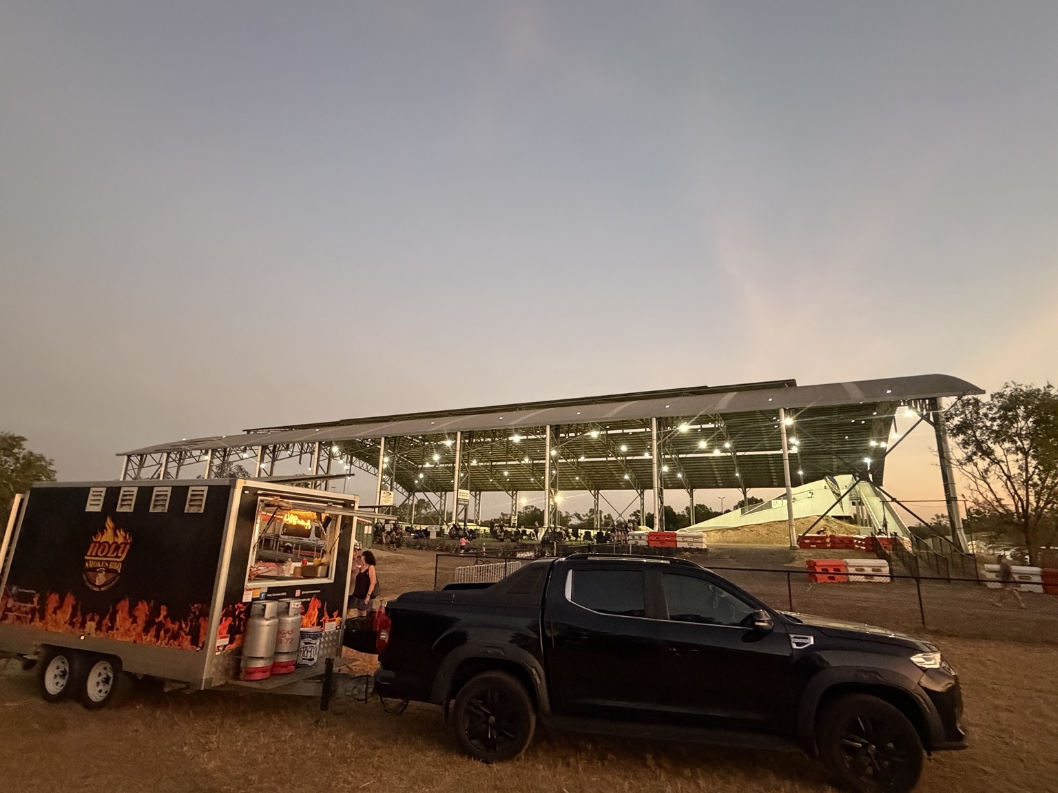 The Holy Smokes BBQ trailer and ute set up at an outdoor event at dusk